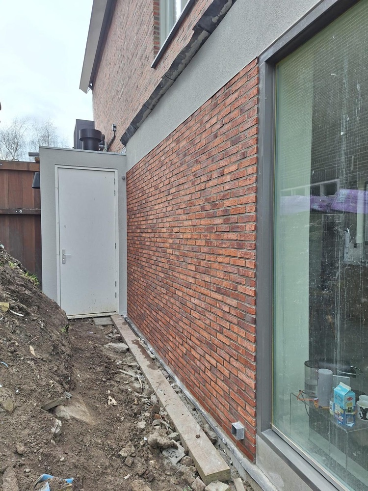 Wand van de aanbouw met steenstrips en voegwerk, naast een grote glazen pui
