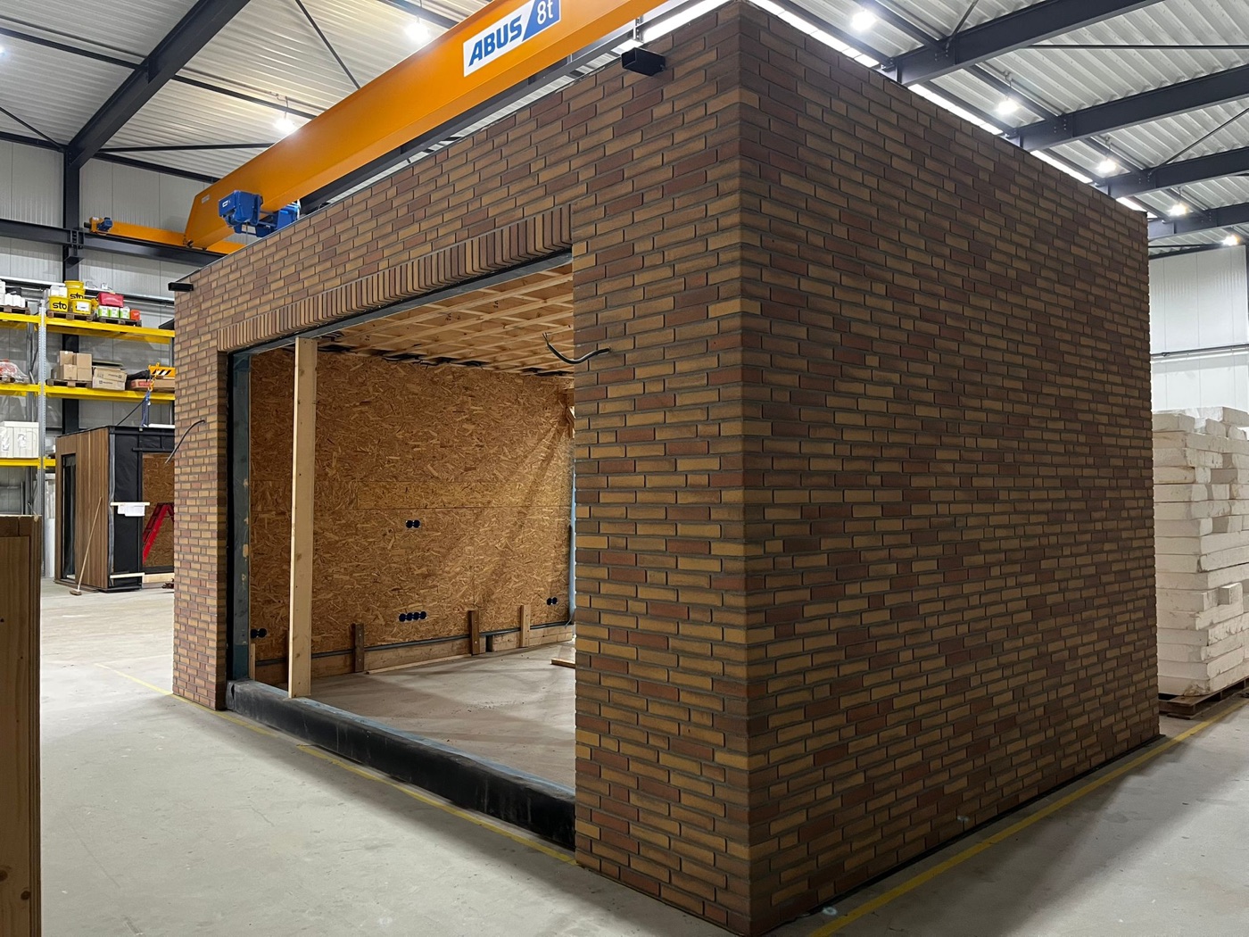 Prefab aanbouw in een werkhal, bekleed met gemêleerd bruine minerale steenstrips, met een grote opening aan de voorzijde