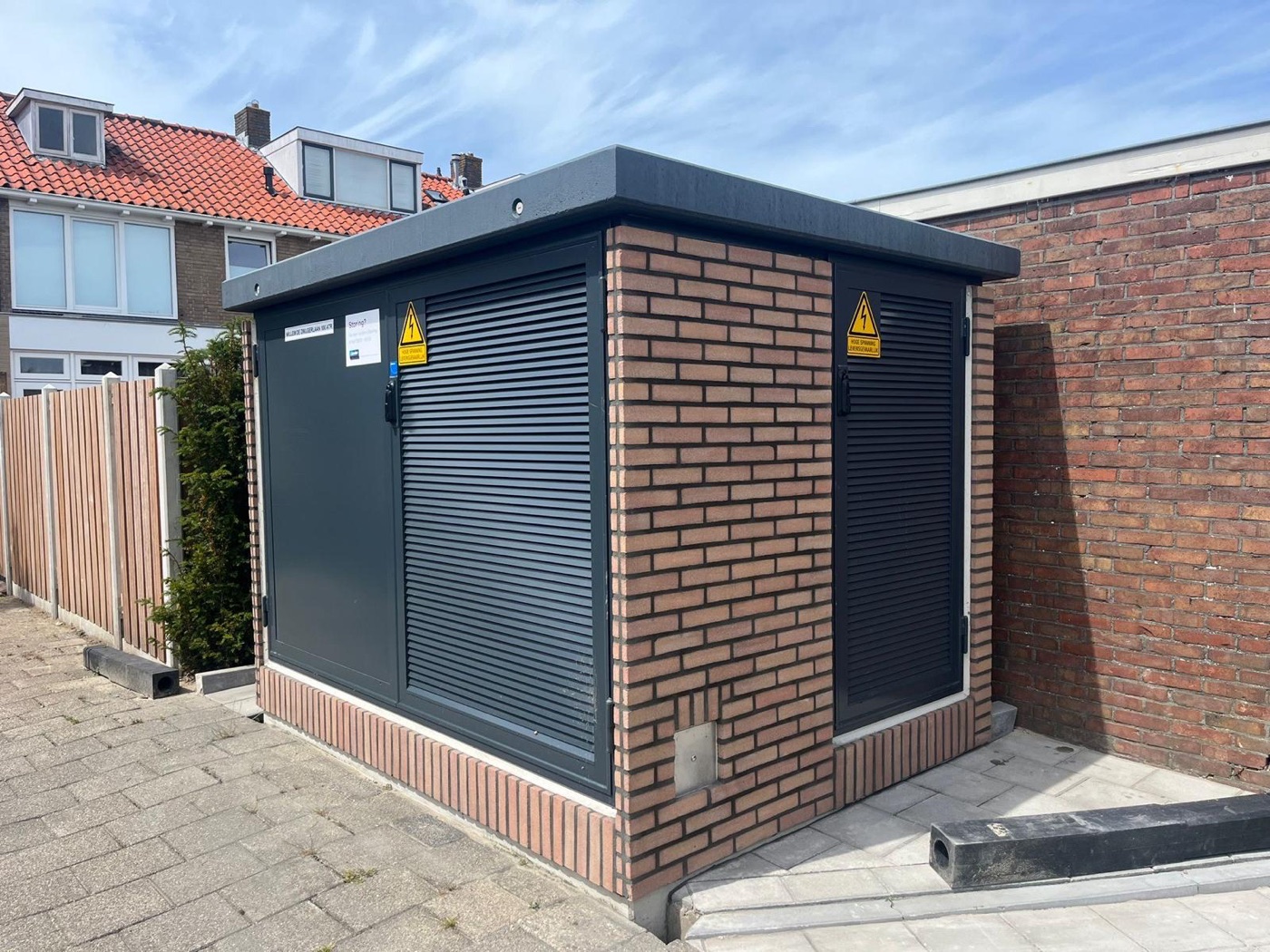 Trafostation met oranjerode steenstrips en antraciet deuren met ventilatieroosters, tussen woningen onder een blauwe lucht