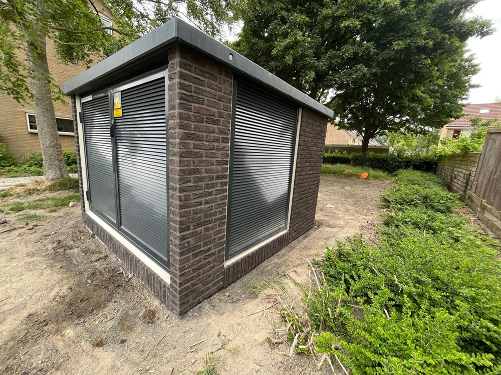 Trafostation met donkerbruine steenstrips en antraciet deuren op een onverhard terrein tussen het groen